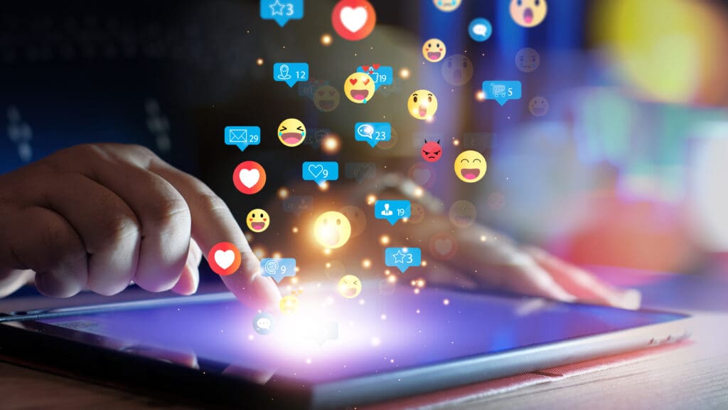 Close-up of a hand interacting with a tablet, surrounded by floating social media icons and emojis, symbolizing online engagement and social media interaction.